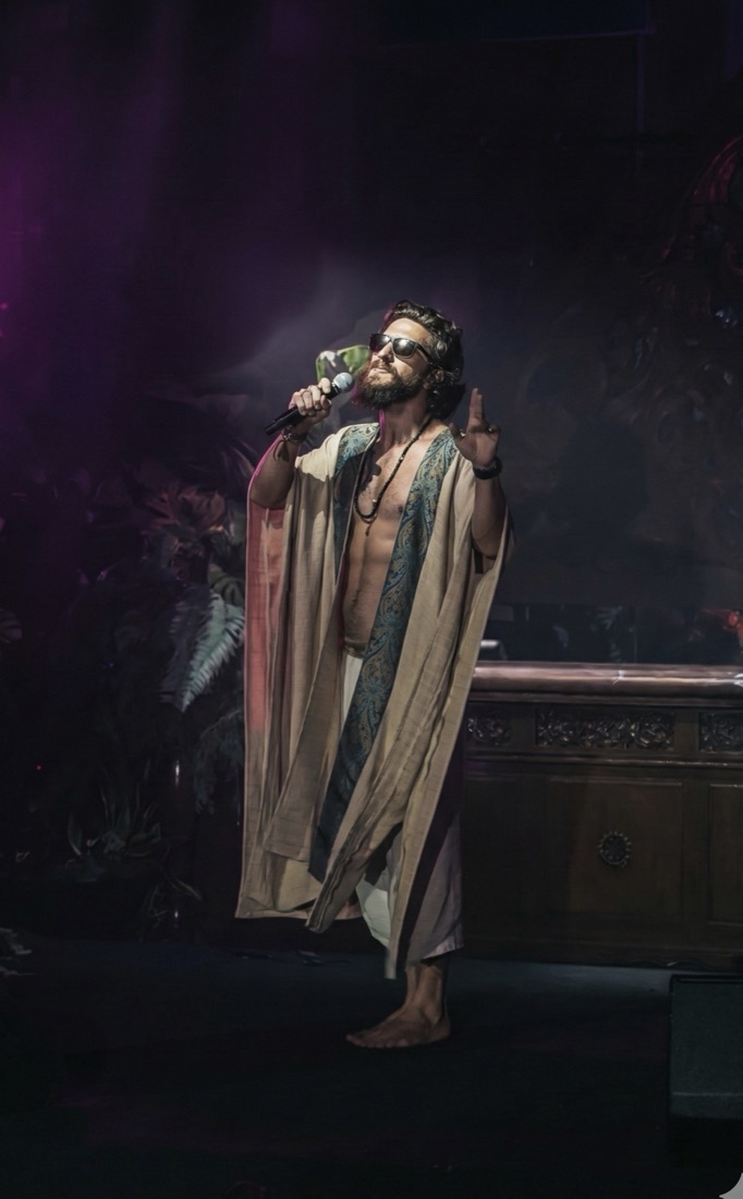 Nikita performing with a microphone on stage in a robe, purple stage haze.
