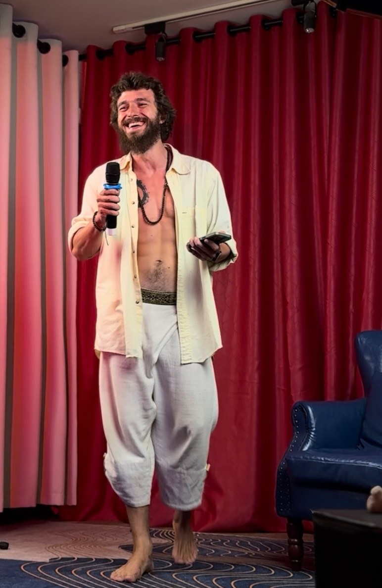 Nikita on stage doing stand-up, mic in hand, open shirt, barefoot, red stage curtain behind.