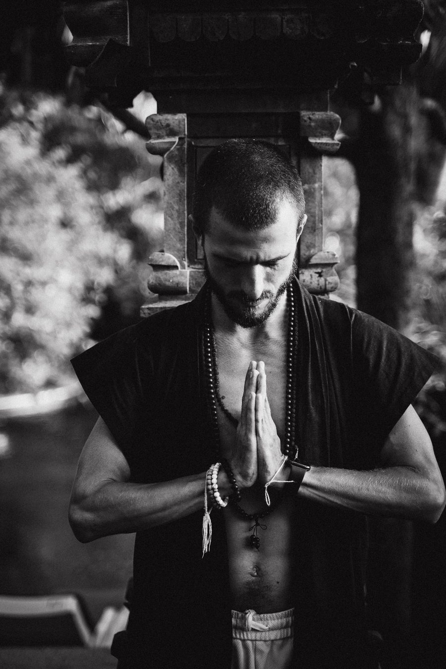 Nikita in prayer, eyes closed, mala beads over a dark robe, before a stone temple shrine.